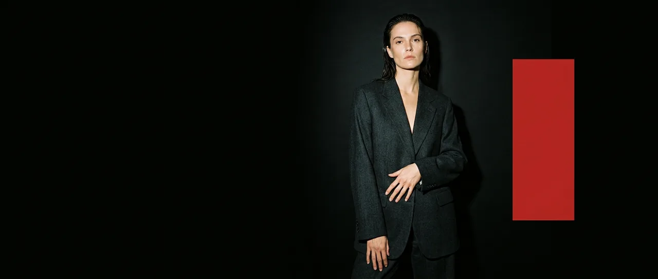 A figure in a dark tailored suit against black, beside a tall red panel.