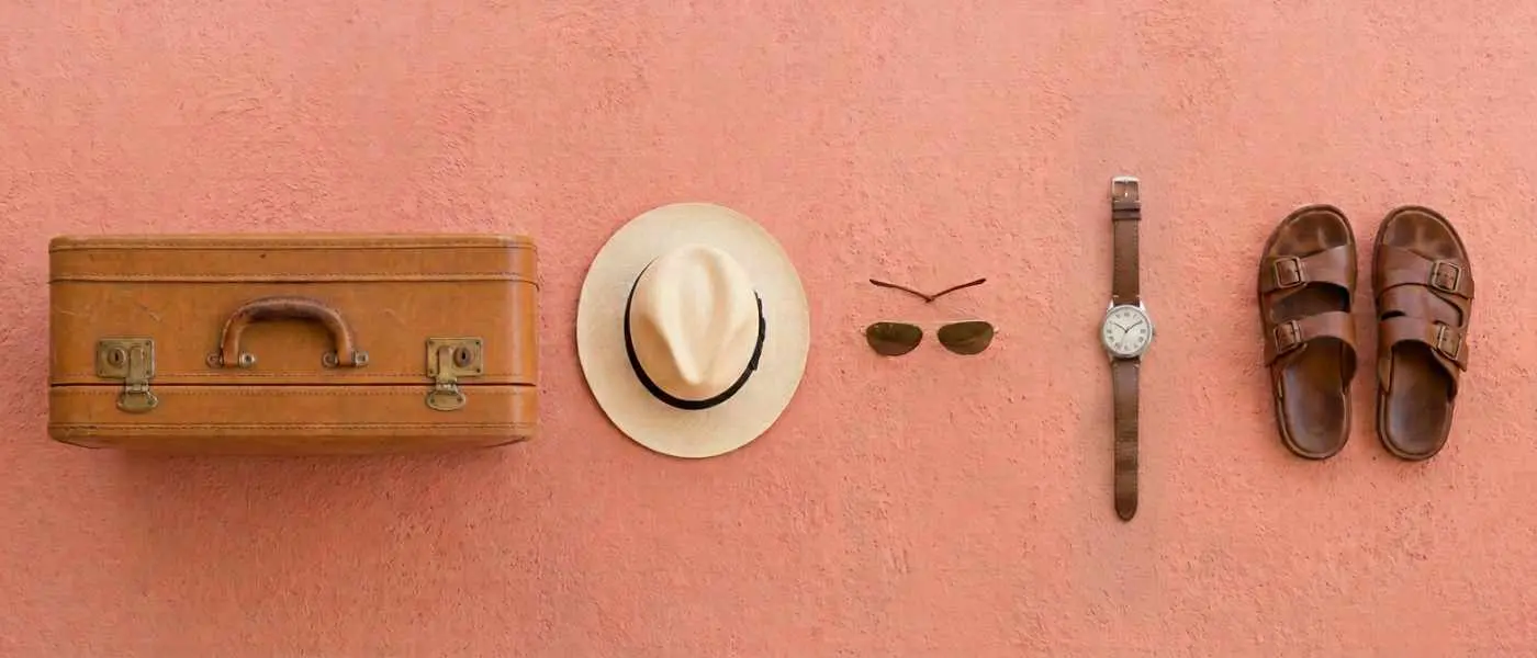 A suitcase, panama hat, sunglasses, watch and sandals laid out on a terracotta wall.